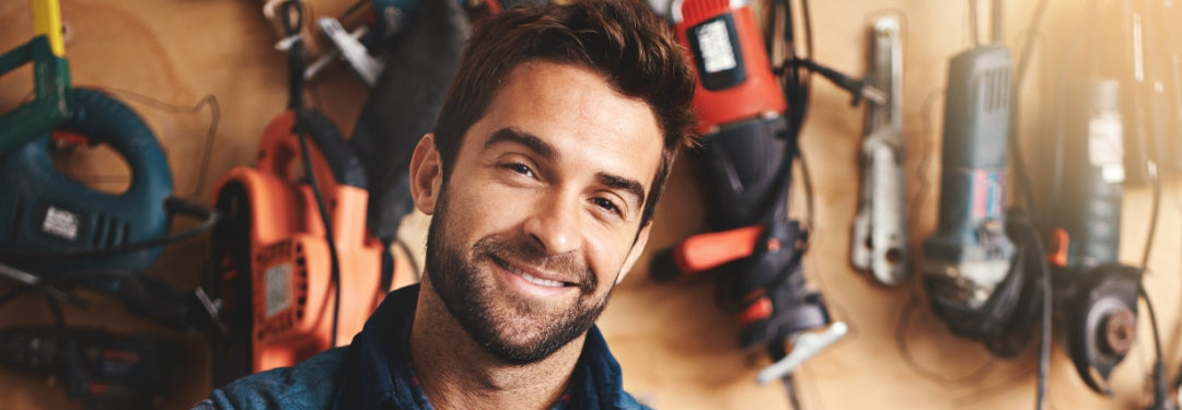 man smiling, standing in workshop with power tools hanging on wall – professional handyman and tool collection background
