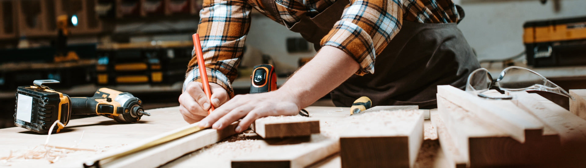 Carpenter measuring and marking wood on workbench with cordless drill and safety glasses – woodworking and construction hand tools in use